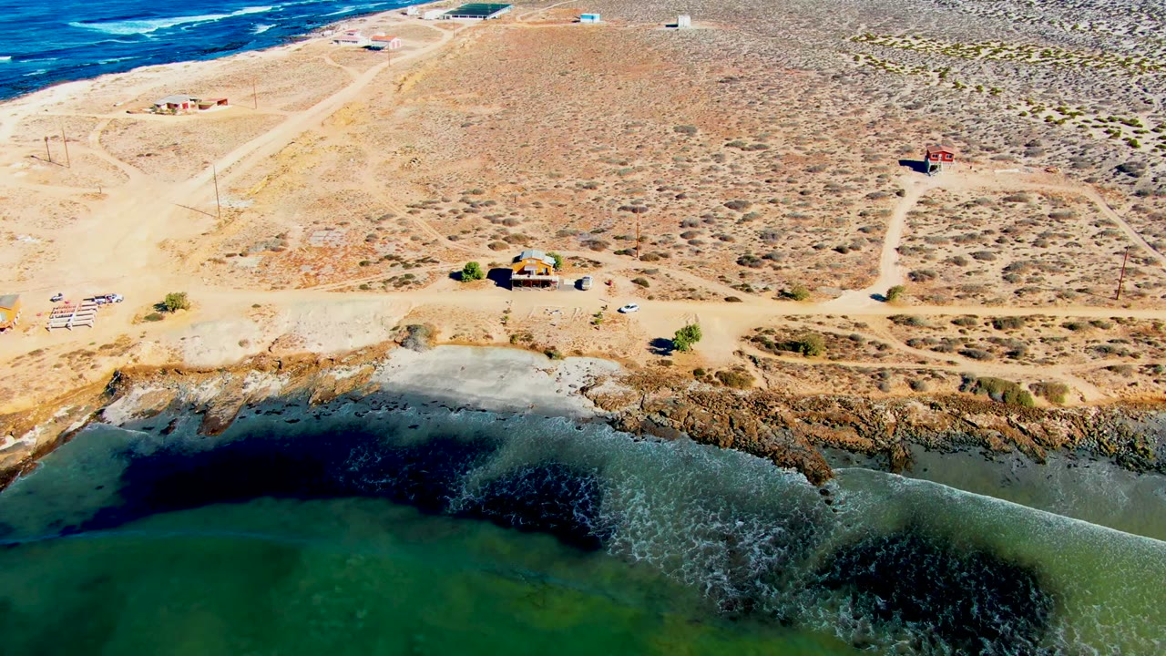 Aerial frame in La Bocana showing the lagoon edge below, a broken rocky shoreline, pale dirt roads crossing dry scrubland, a few vehicles, small structures, ocean near the horizon, and no visible people.