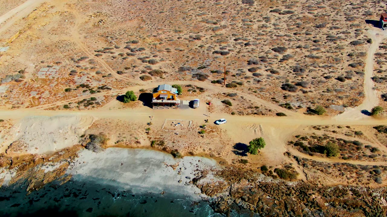 Overhead view in La Bocana, Baja California Sur, showing a small house beside a dirt road, sparse desert scrub, a white car, and shoreline water at the lower edge, with no visible people.