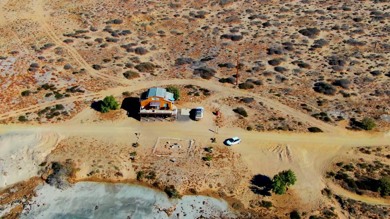 Top-down La Bocana view showing a dirt road running past a small house, a white car near the road, dry scrubland above, and blue-green water cropped along the bottom, with no visible people.