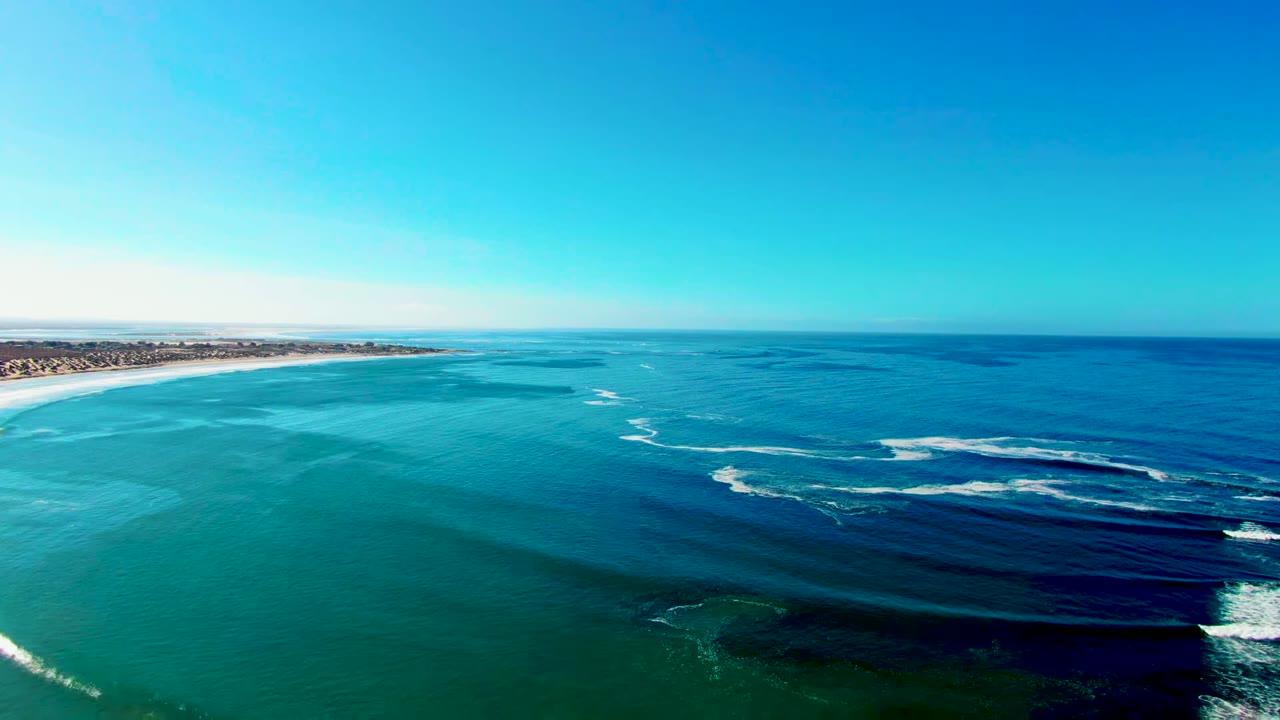 Opening aerial frame over La Bocana, Baja California Sur, showing turquoise water, darker offshore bands, white surf, a sandy shoreline at left, distant coastal buildings, and no visible people.