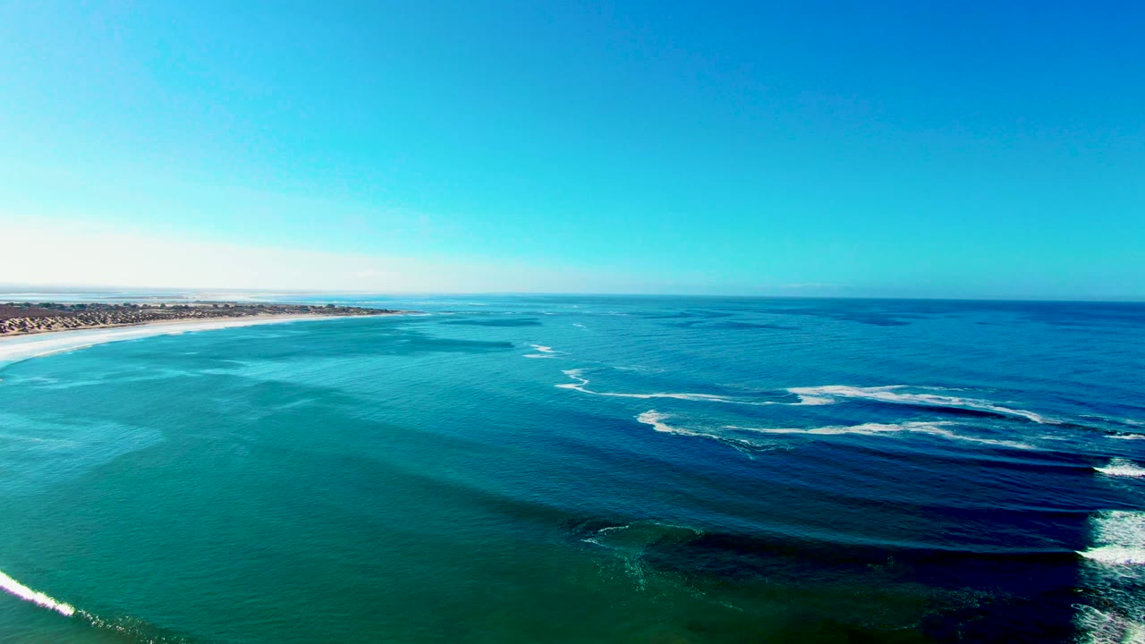 Wide aerial coastline with white surf crossing blue-green water.