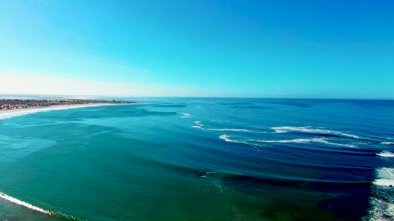 Aerial view of La Bocana at 0:02, with pale turquoise shallows near the beach, darker water patches offshore, foamy wave crests in the midground, and no visible people.