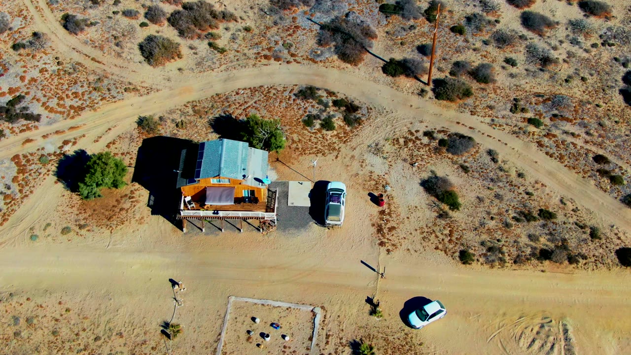 Overhead drone frame in La Bocana, Baja California Sur showing a small shack with a pale metal roof, a parked light-colored vehicle, sandy roads, scrub vegetation, and tiny visible people near the structure.