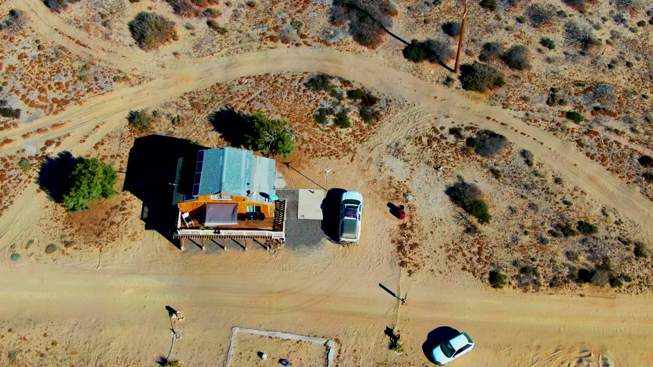 Aerial view of the shack, parked vehicle, scrubland, and a light-colored car on the dirt road.