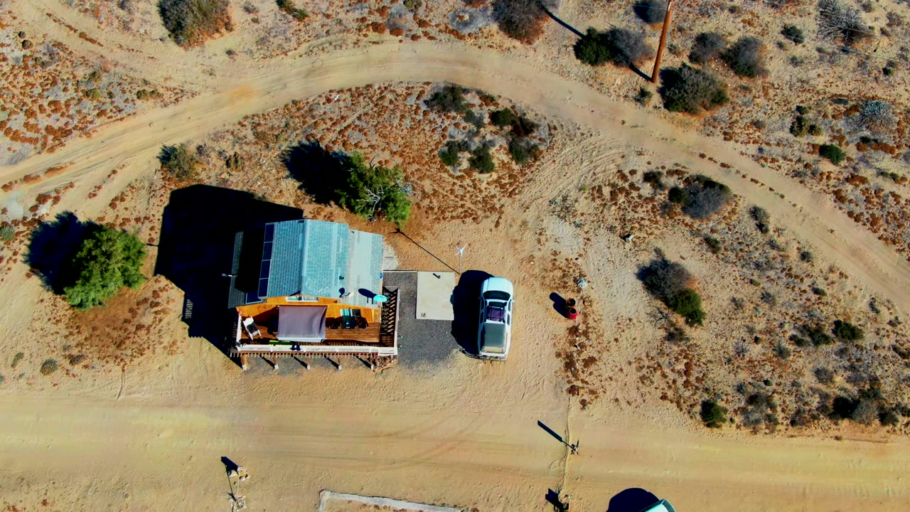 Top-down frame in La Bocana, Baja California Sur showing the shack and parked vehicle at right of the structure, winding dirt tracks above, dry scrubland, strong sun shadows, and tiny visible people near the road.