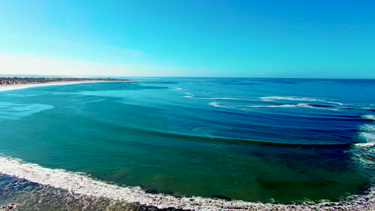 Aerial view at La Bocana, Baja California Sur, with blue ocean, turquoise shallows, white surf near the lower edge, distant coastline at left, and no visible people.