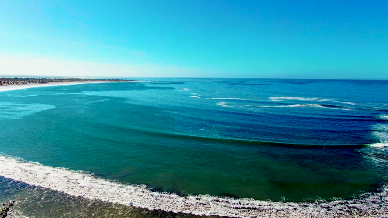 Wide drone view of turquoise water and white foam along the beach edge.