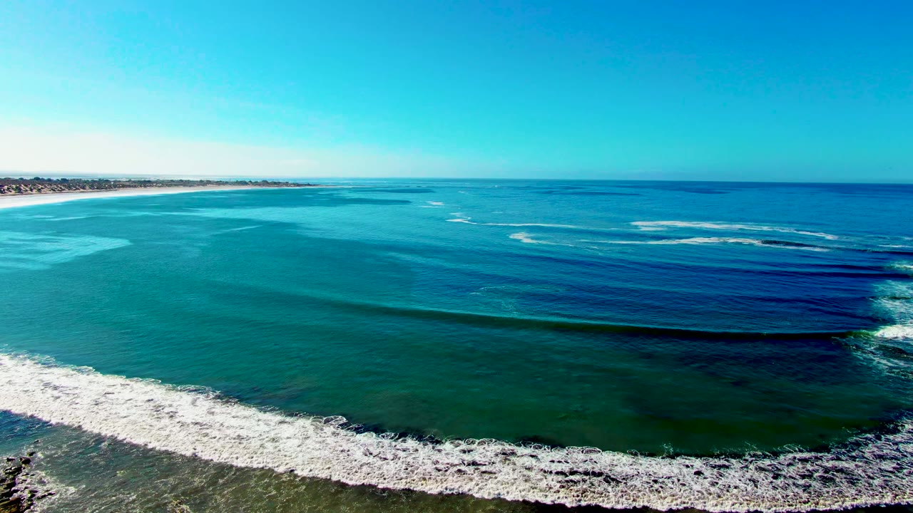 High aerial frame at La Bocana, Baja California Sur, with parallel dark swell lines moving through blue-green water toward a bright foam strip and rocky lower edge, with no visible people.