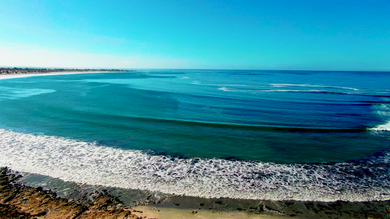 Aerial frame over La Bocana, Baja California Sur showing turquoise water, a white surf band, sandy beach, rocky cliff foreground, clear sky, and no visible people.