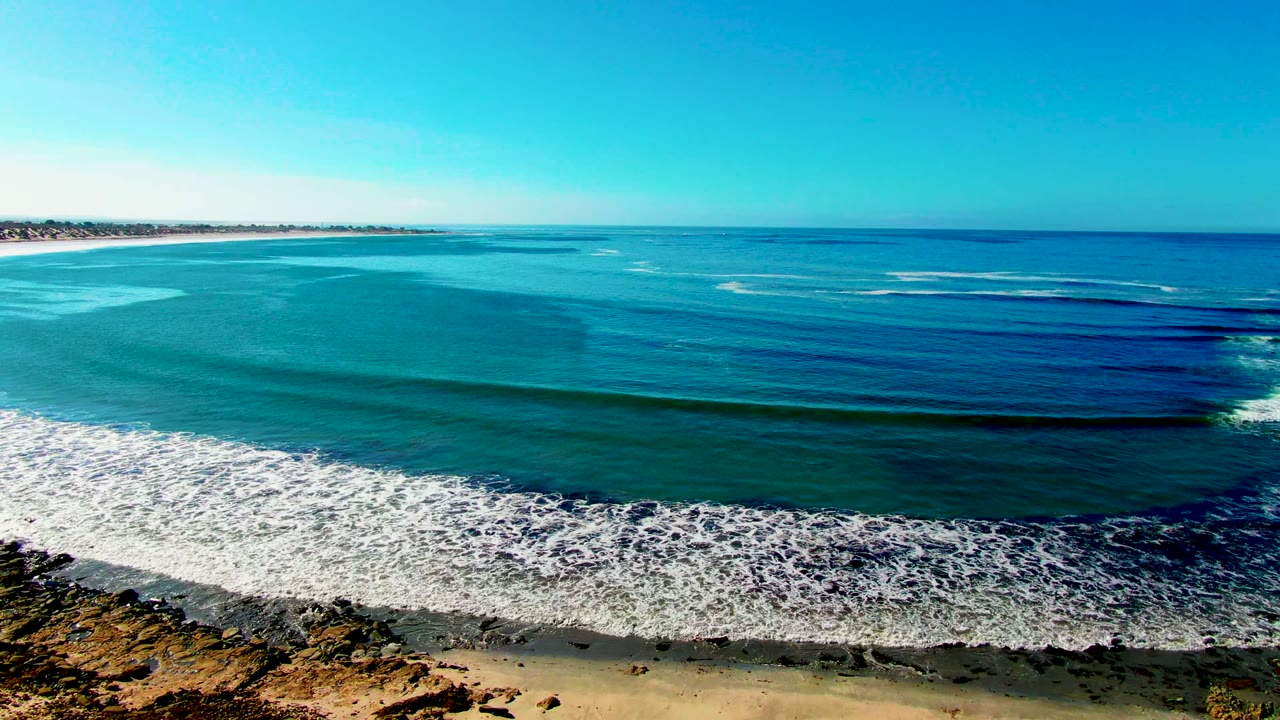 White surf spreads across shallow blue-green water beside a sandy beach and rocky foreground.