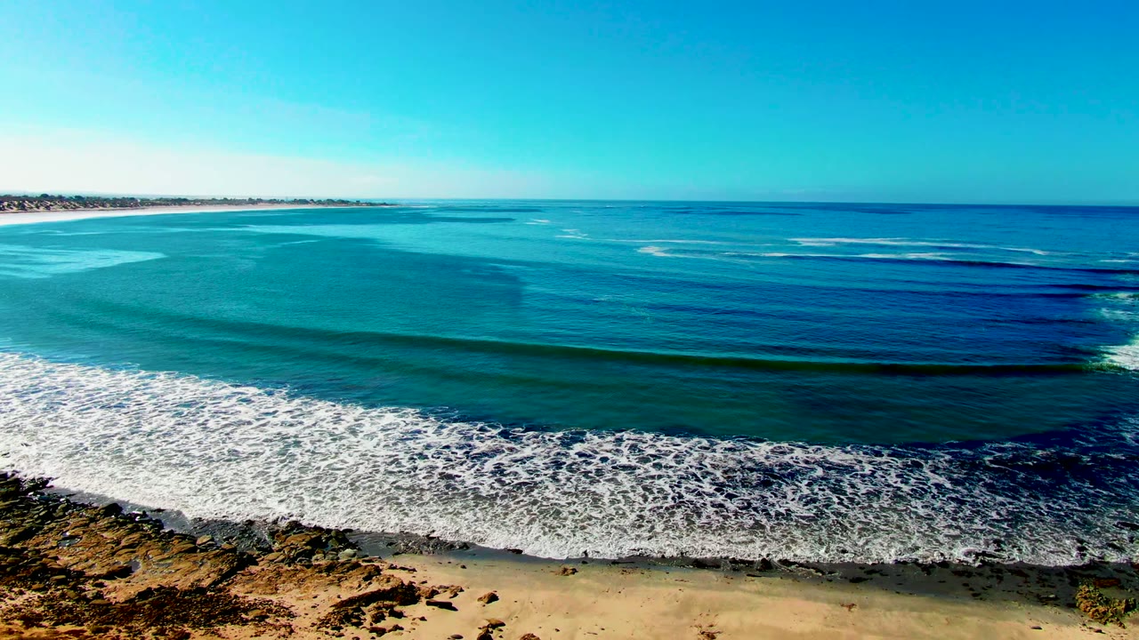 La Bocana coastline from above, with layered deep blue and turquoise water, a breaking surf band near the lower frame, sandy shore, rocky bluff edge, and no visible people.
