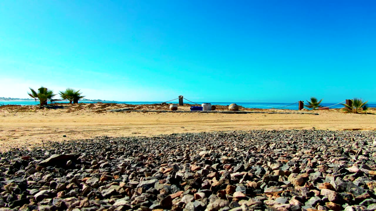 Opening frame of a La Bocana pebbled beach with a rocky foreground, sandy middle strip, small palms, ocean horizon, clear blue sky, and a tiny distant figure near the shore.
