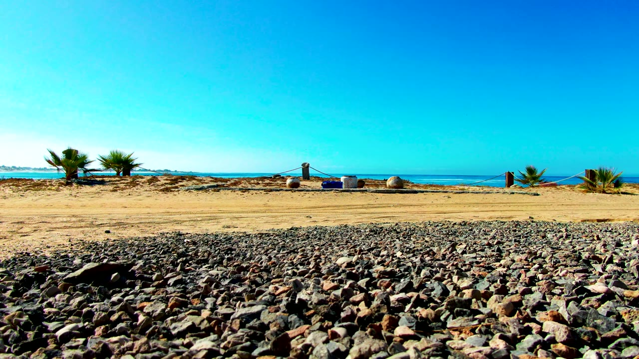 A wide La Bocana shoreline frame holds pebbles across the lower image, a flat sandy band, scattered palms, the blue ocean, and a small distant person near the sandy shore.