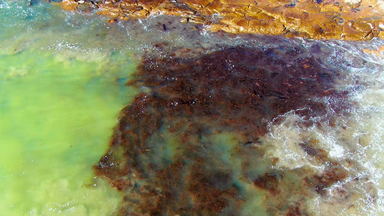 At two seconds, the overhead La Bocana frame includes ochre rock along the upper edge, green water below, and a broad dark algae or sediment patch edged with foam, with no visible people.