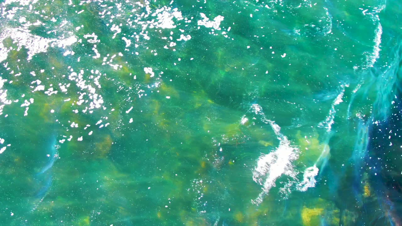 An overhead view in La Bocana, Baja California Sur shows rippled clear water, mottled seaweed, pale rocks, and scattered white highlights with no visible people.