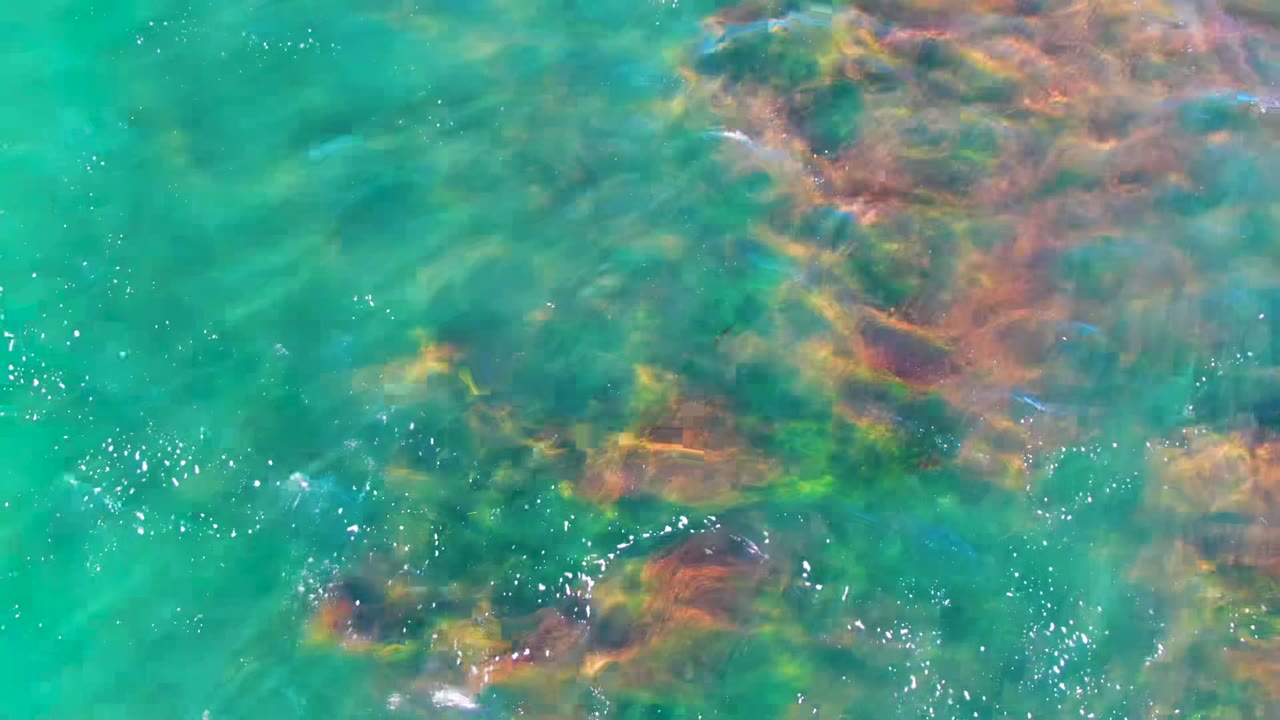 Overhead view of clear turquoise water at La Bocana, Baja California Sur, with submerged brown and green rocks visible beneath light ripples and no visible people.