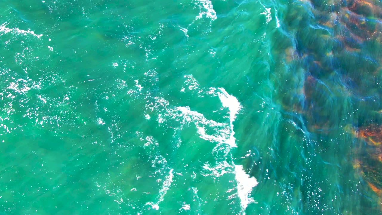Overhead coastal water view in La Bocana, Baja California Sur, with reddish brown and green rocks showing through rippled turquoise water and no visible people.