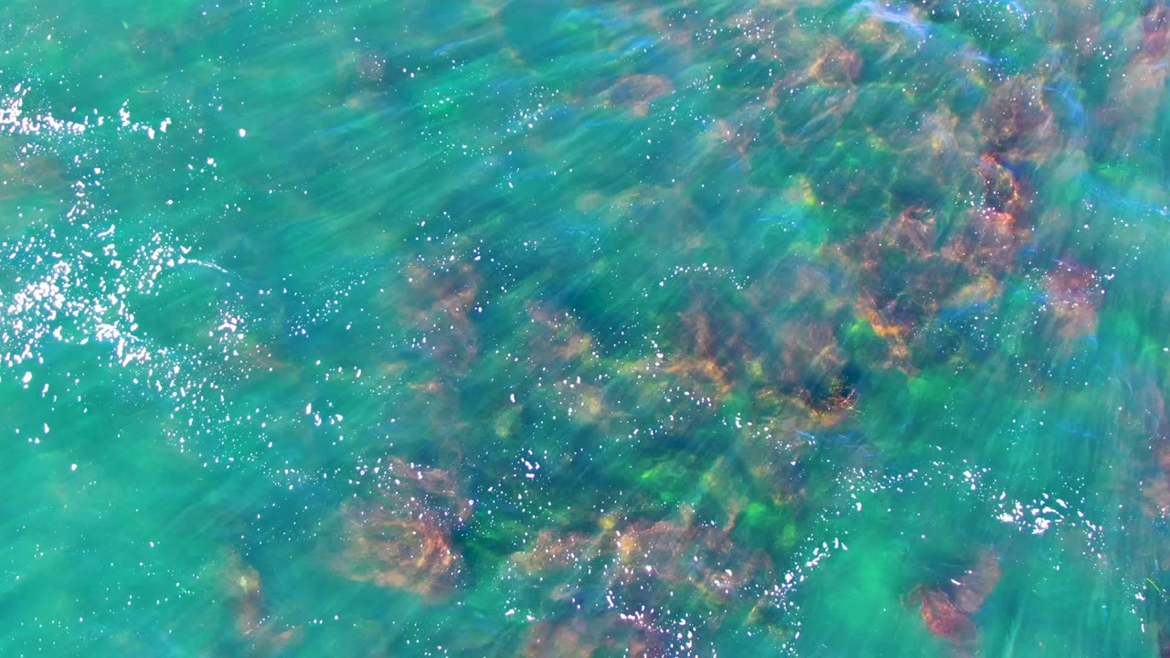 Overhead view in La Bocana, Mexico, where clear teal water reveals clustered underwater rocks with slightly shifted ripples and no visible people.