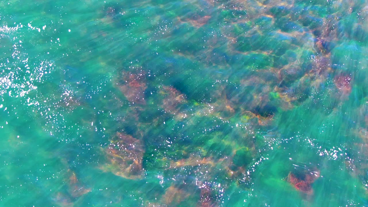 A later overhead frame from La Bocana, Mexico, with the rocky seabed more spread across the upper frame, bright water highlights, and no visible people.