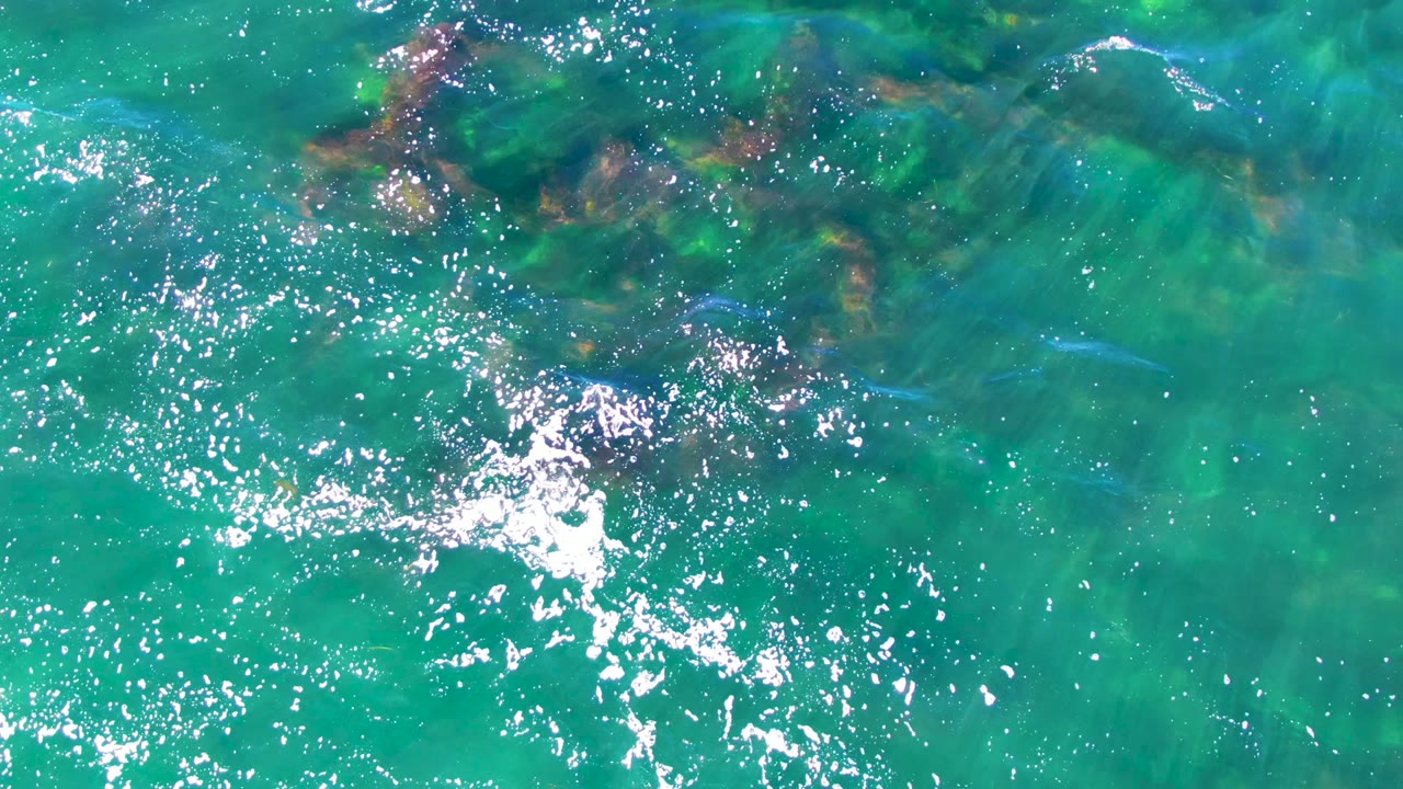 Overhead view of clear turquoise water in La Bocana, Baja California Sur, with submerged rocky patches near the upper frame and no visible people.
