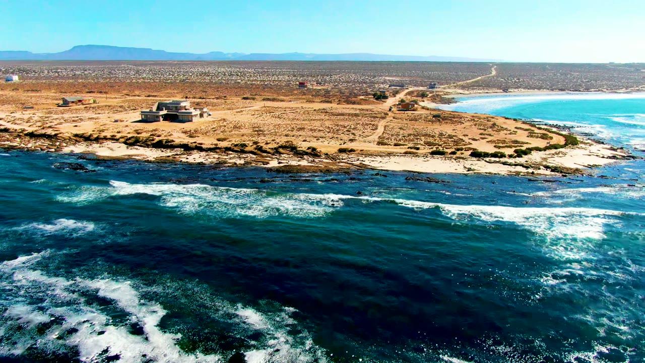 Opening aerial frame over La Bocana, Baja California Sur, with dark blue ocean foam in front, a sandy point and small building beyond it, distant mountains, and no visible people.