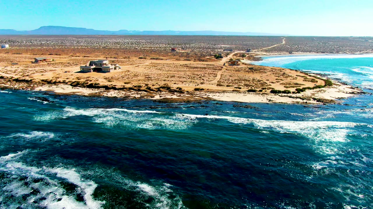 Wide drone view of white surf breaking below a dry shoreline and small structure.