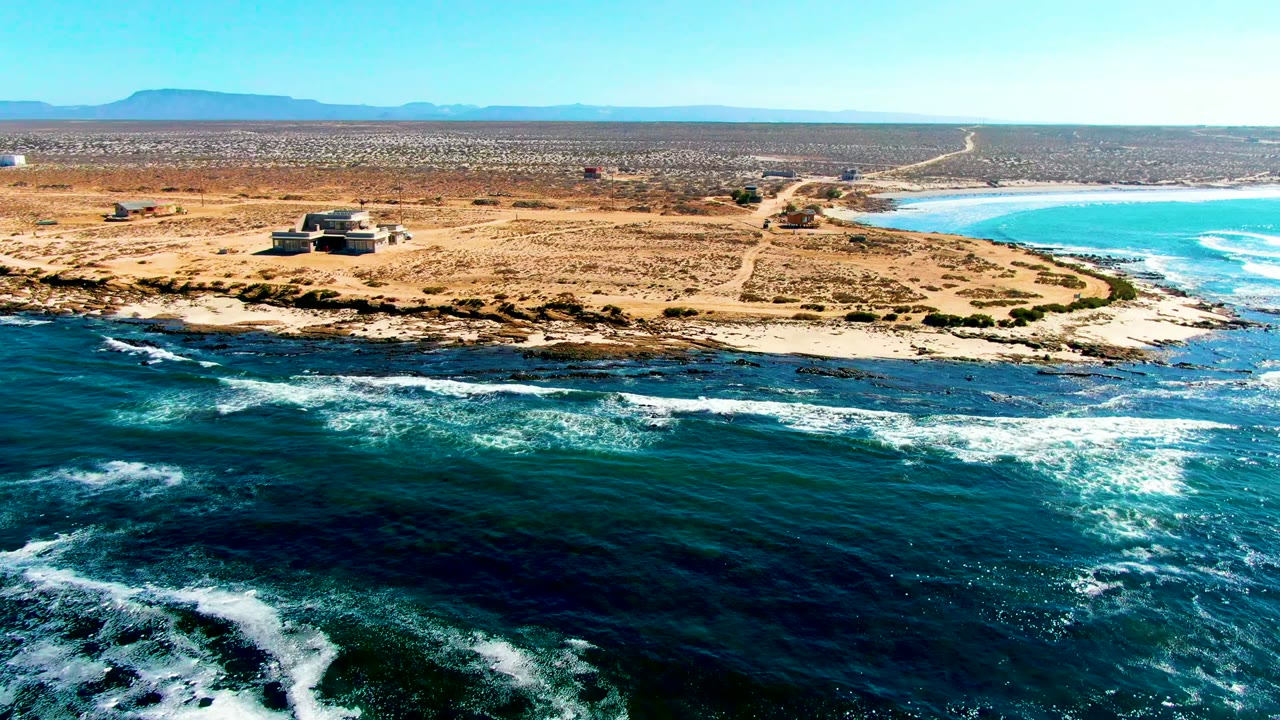 Aerial frame at 0:02 with foam bands crossing the ocean foreground, a dry La Bocana coastal point with a small structure, pale dirt roads, mountains on the horizon, and no visible people.