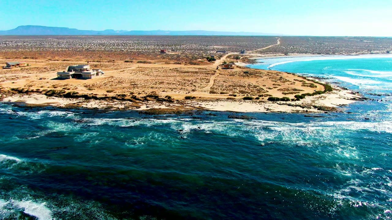 Aerial frame over La Bocana, Baja California Sur with dark teal surf in the foreground, a sandy headland and small building on the left, distant desert mountains, and no visible people.