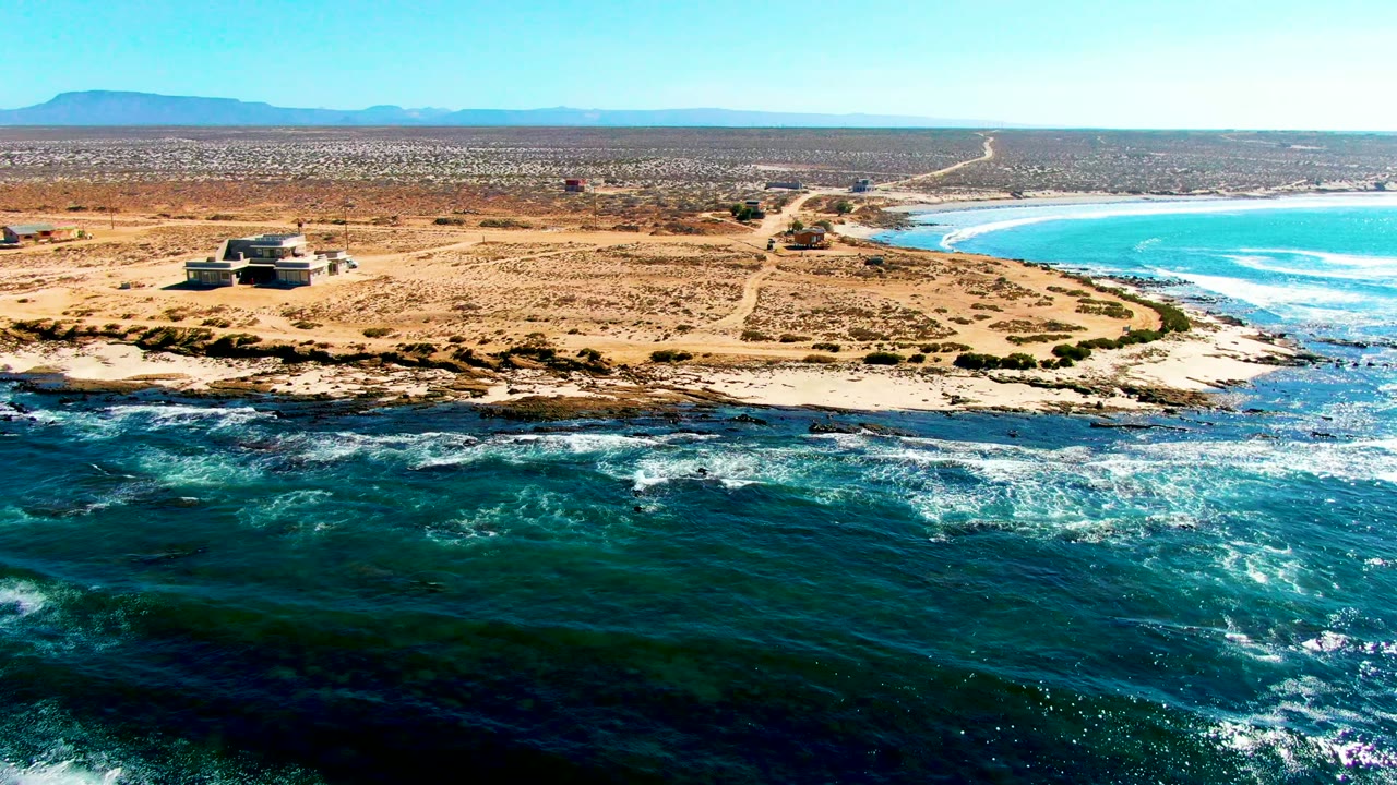 Wide aerial coastline with waves breaking below a sandy point and small structure.