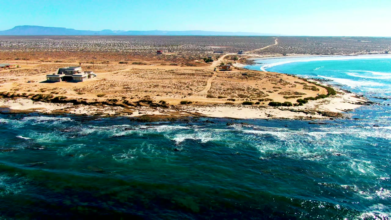 La Bocana aerial frame with the headland building left of center, a pale dirt road running into the desert, turquoise water along the right edge, white surf below, and no visible people.