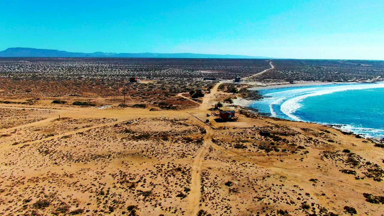 Wide aerial frame over La Bocana, Baja California Sur, with sandy scrubland, a dirt road, small coastal buildings, blue ocean waves at right, distant hills, and no visible people.