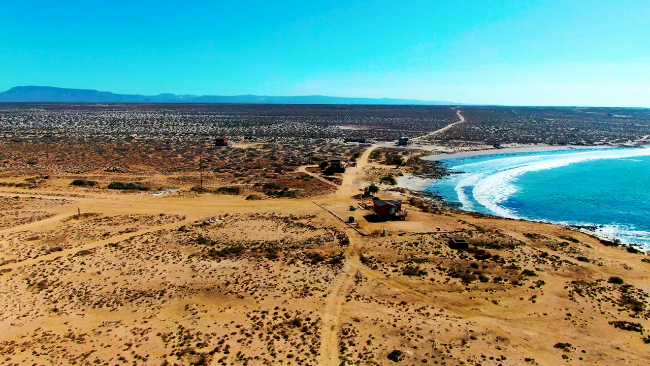 High aerial view of desert scrub, dirt tracks, small buildings, and surf along the right edge.