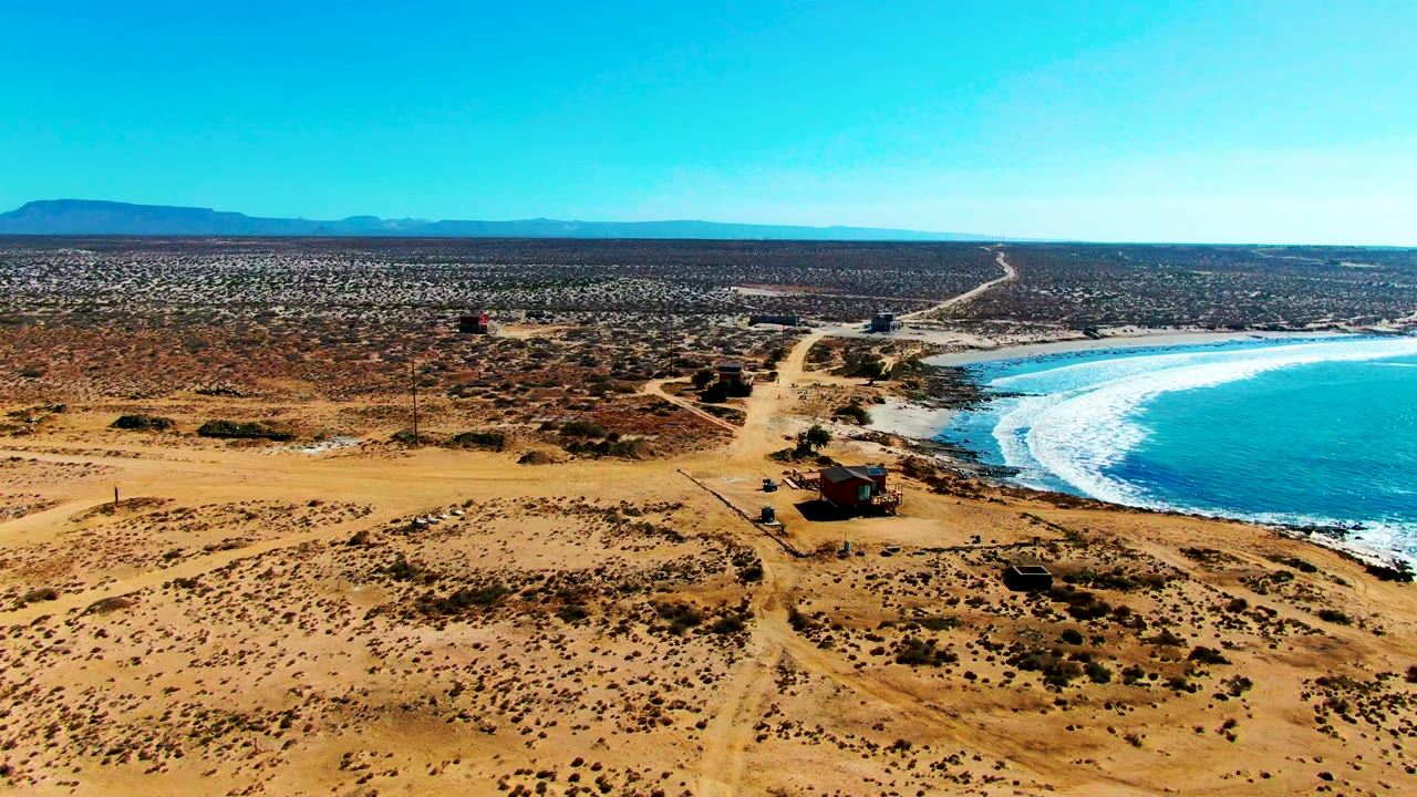 La Bocana aerial frame with more of the beach and turquoise water visible at right, the dirt road cutting through dry terrain, scattered buildings near the road, distant low hills, and no visible people.