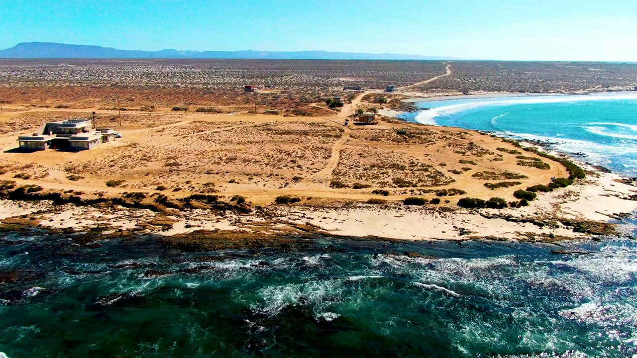 Aerial frame over La Bocana, Baja California Sur, showing tan sandy ground with sparse scrub, small coastal buildings at left, blue ocean at right, rocky surf in front, and no visible people.