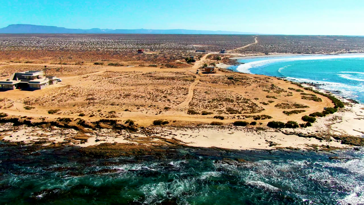 Aerial frame over La Bocana with dirt tracks through scrubby sand, a few structures near the coast, turquoise water curving along the right edge, distant mountains, and no visible people.