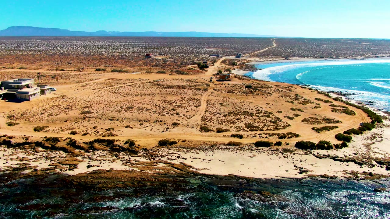 Aerial frame at La Bocana showing a sandy coastal bluff with scattered scrub, small buildings near the left and center, bright ocean on the right, white wave bands, and no visible people.