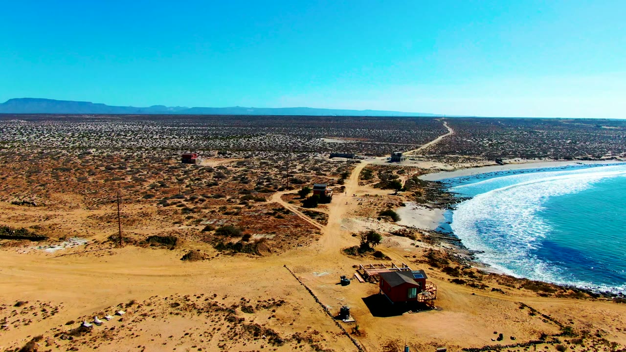 Aerial view over La Bocana, Baja California Sur, with a sandy road, small huts, scrub desert, turquoise surf on the right, distant mountains, and no visible people.