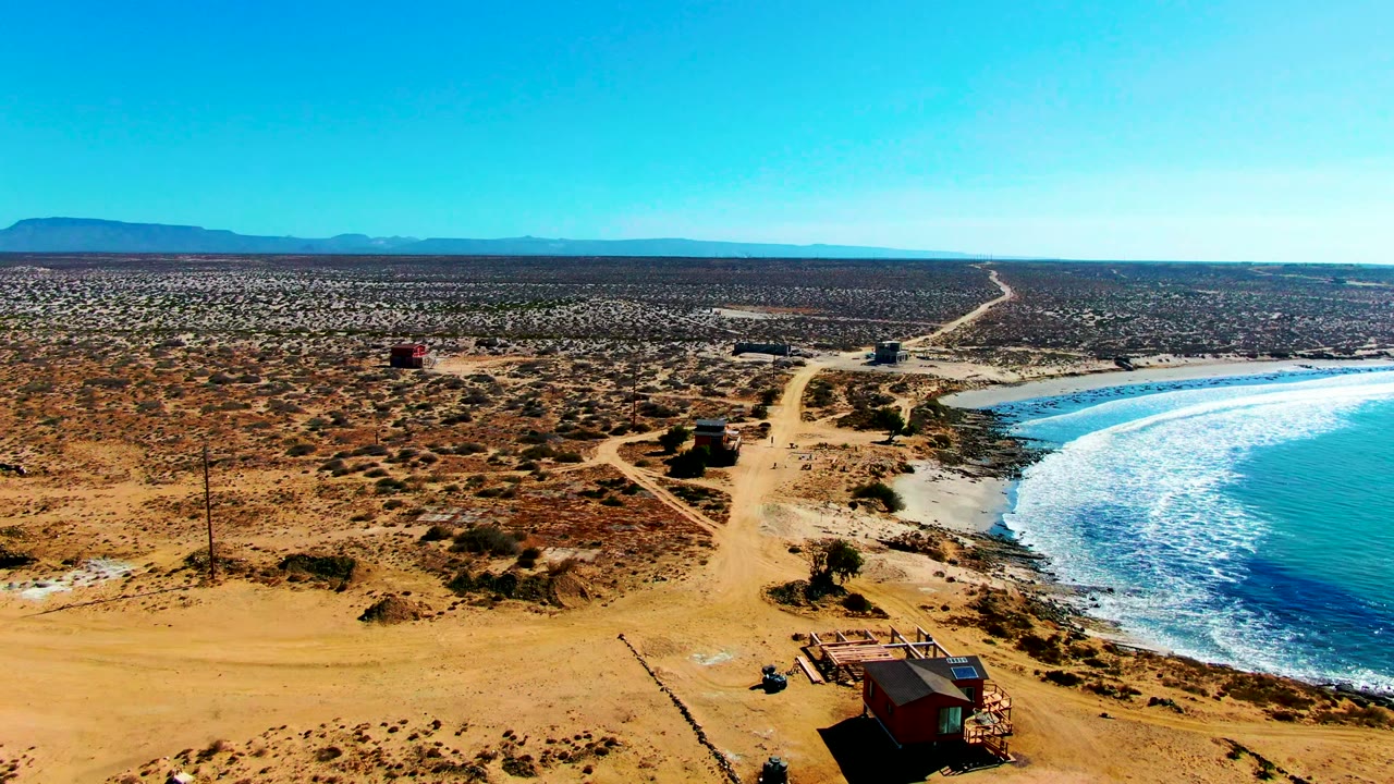 Drone view of sandy tracks, small coastal huts, desert scrub, and blue ocean surf.