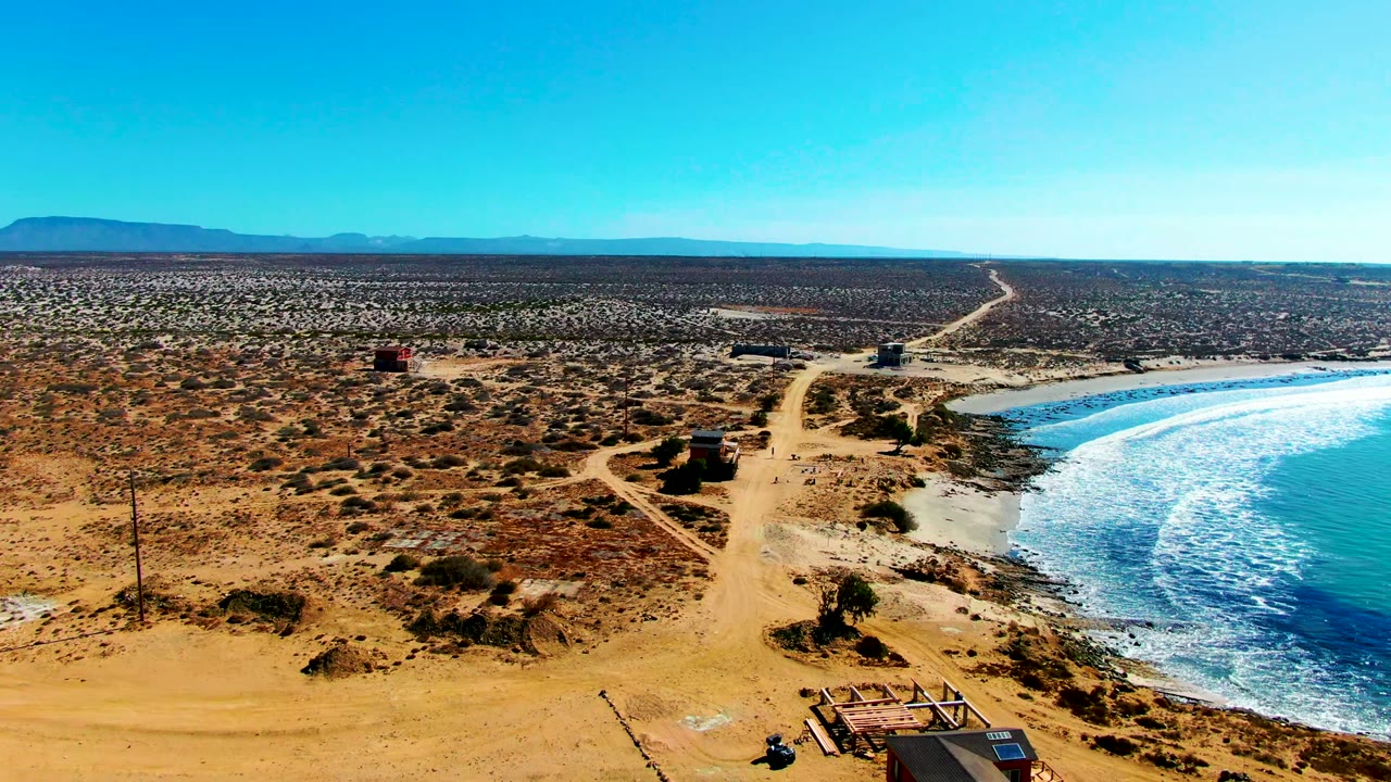 La Bocana coastal desert appears from above with a small hut near the bottom edge, branching sandy tracks, a bright surf line on the right, open scrubland inland, and no visible people.