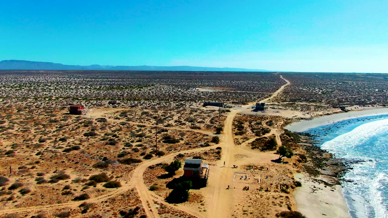 Aerial view over La Bocana, Baja California Sur, with a sandy dirt road running past desert scrub toward blue ocean on the right and no recognizable people visible.