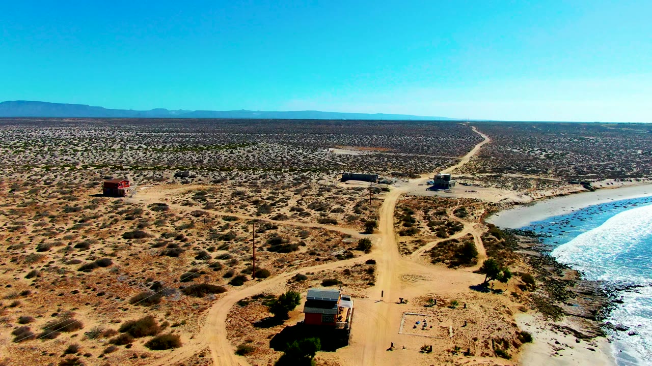 Elevated frame in La Bocana showing the dirt road centered through arid scrub, a small vehicle or trailer near the roadside, bright surf at right, and no recognizable people visible.