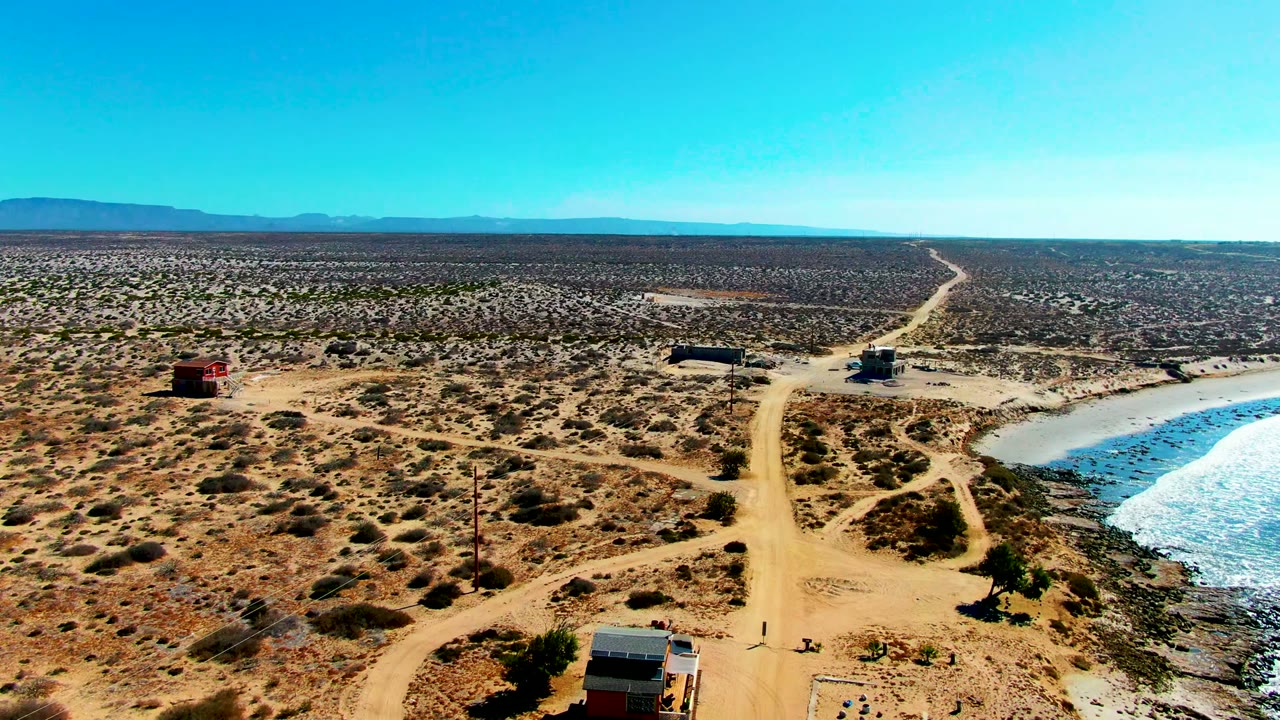 Aerial view in La Bocana with the road bending near small structures, desert brush spreading to the horizon, blue water along the right edge, and no recognizable people visible.