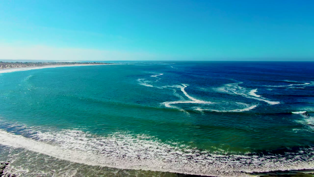 Aerial frame over La Bocana, Baja California Sur showing turquoise surf bands, deep blue water offshore, sandy beach at the bottom edge, and no visible people.