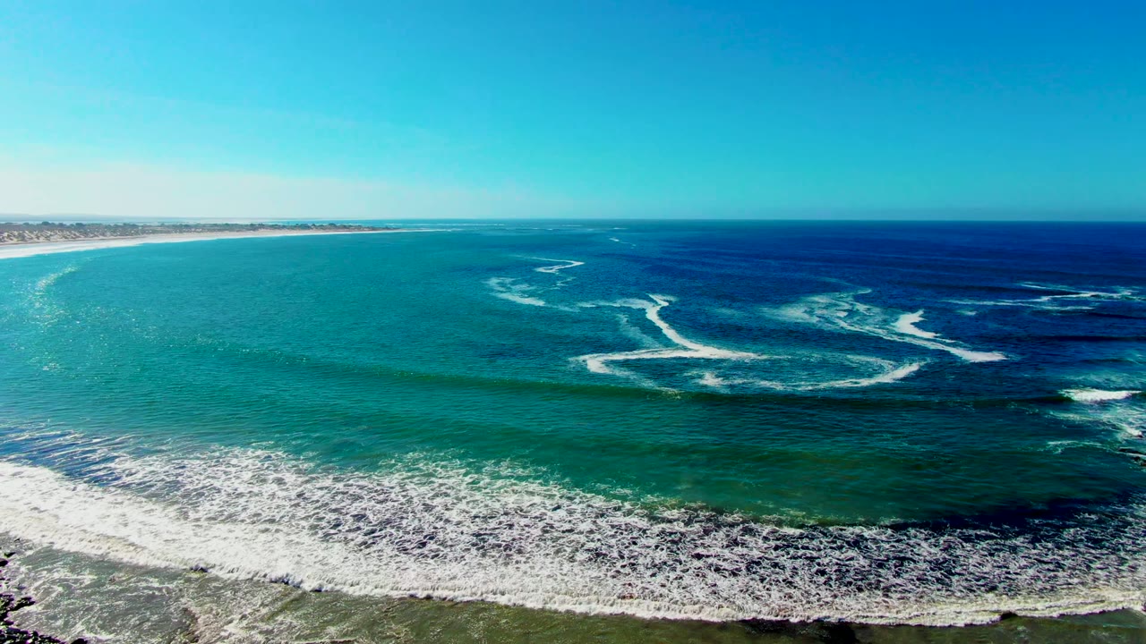 Aerial frame over La Bocana, Baja California Sur showing broken white wave streaks across the midground, sandy coast along the lower edge, and no visible people.