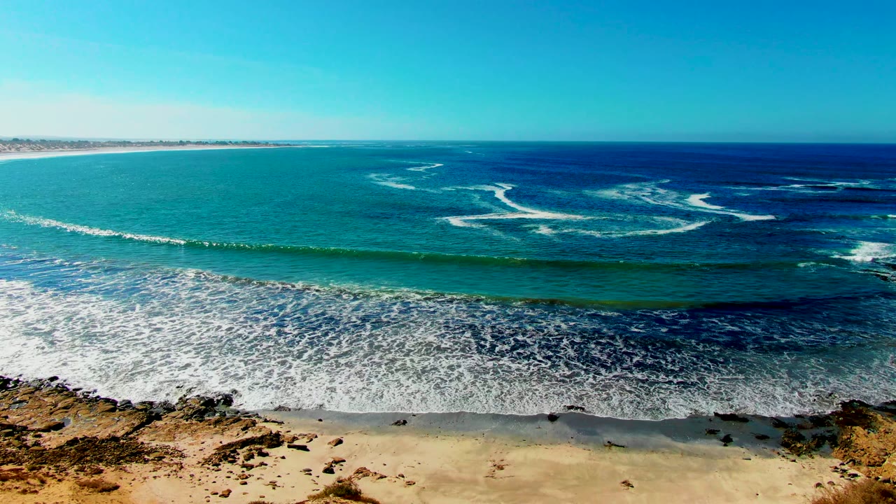 Elevated view over La Bocana, Baja California Sur, with turquoise water, white surf lines, sandy beach, rocky shoreline patches, and no visible people at the start of the clip.