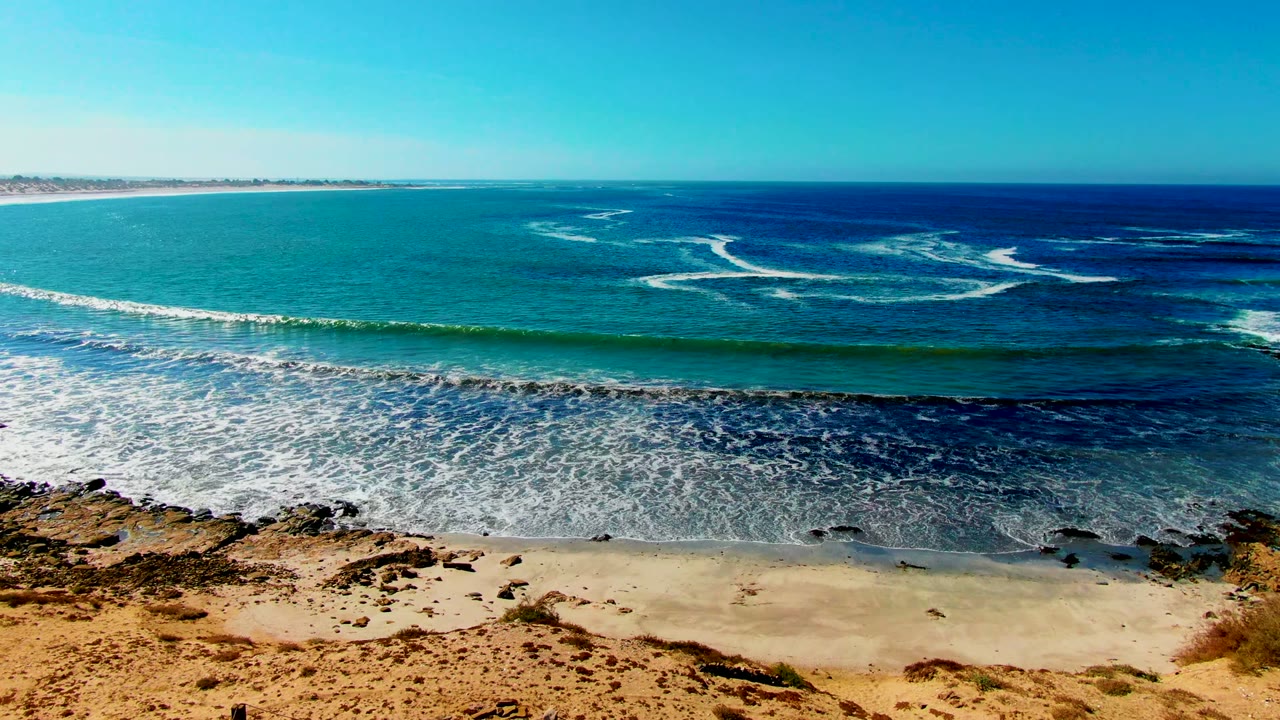 At 0:02, a green-blue swell line approaches the sand at La Bocana while foam gathers in the shallows and rocky outcrops mark the lower edge, with no visible people.