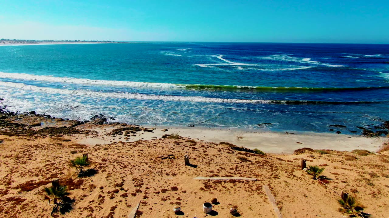 Early frame of La Bocana beach showing tan sand, low plants, white surf lines, blue ocean, clear sky, and no visible people.