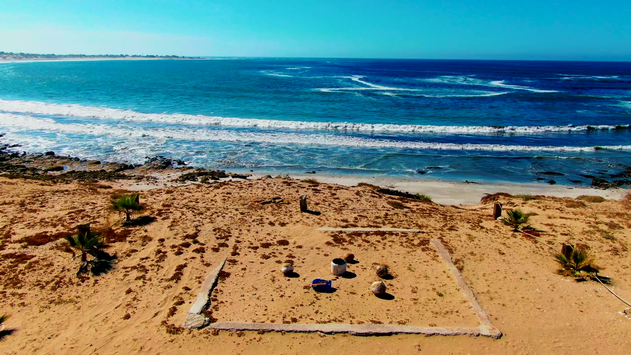 La Bocana beach view with bright surf, dark blue water farther out, sand tracks, sparse vegetation, and no visible people as the pan shifts along the shore.