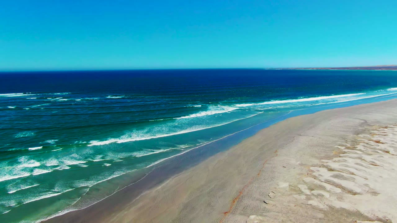 At La Bocana in Baja California Sur, an aerial view shows turquoise surf approaching a pale sandy beach under a clear blue sky, with no visible people.