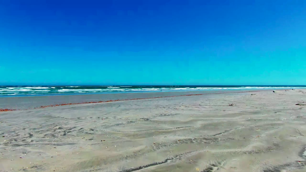 Wide aerial frame over La Bocana beach with pale rippled sand in front, white surf bands along the ocean, a clear blue sky, and no visible people.